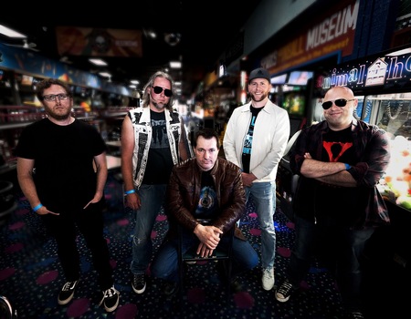 The members of Vultures of Culture standing in a dark bowling alley with Luke sitting in a chair in the center.